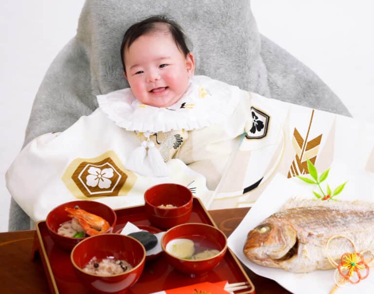 一生食べ物に困らないようにと願う、尾頭付きの鯛や赤飯の御膳を前に、白い祝着を纏ってにっこりと微笑む赤ちゃん